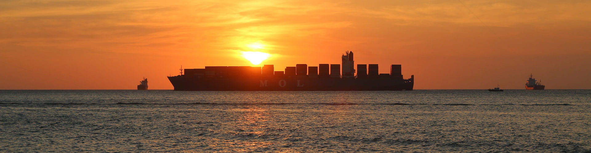 Large cargo ship in the ocean with three small boats nearby, with orange sky and sun setting.